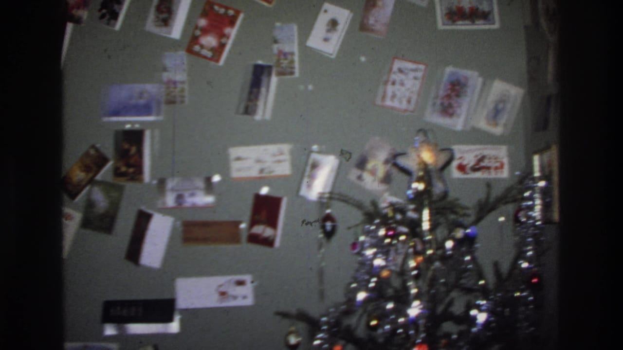 Christmas cards still on wall as camera pans right; top of a decorated pine tree with lights and tinsel enters frame.