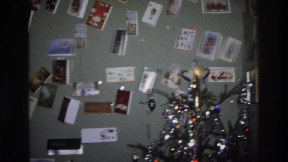 Mid-pan frame showing the Christmas card wall and the emerging top of a tinsel-decorated tree with string lights, from a 1975 Washington DC home movie.