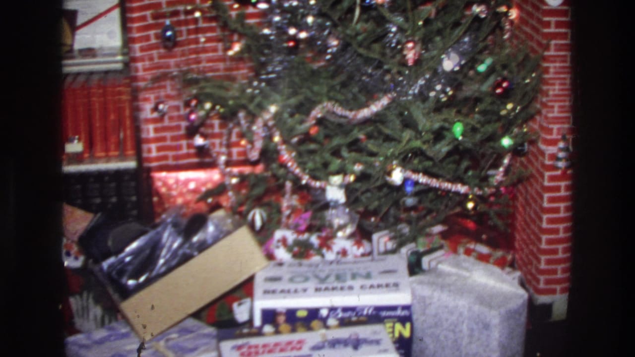 Closer view of a 1975 decorated Christmas tree with string lights and ornaments; wrapped presents at the base include a cardboard box with legible printed branding, set before a red brick fireplace.