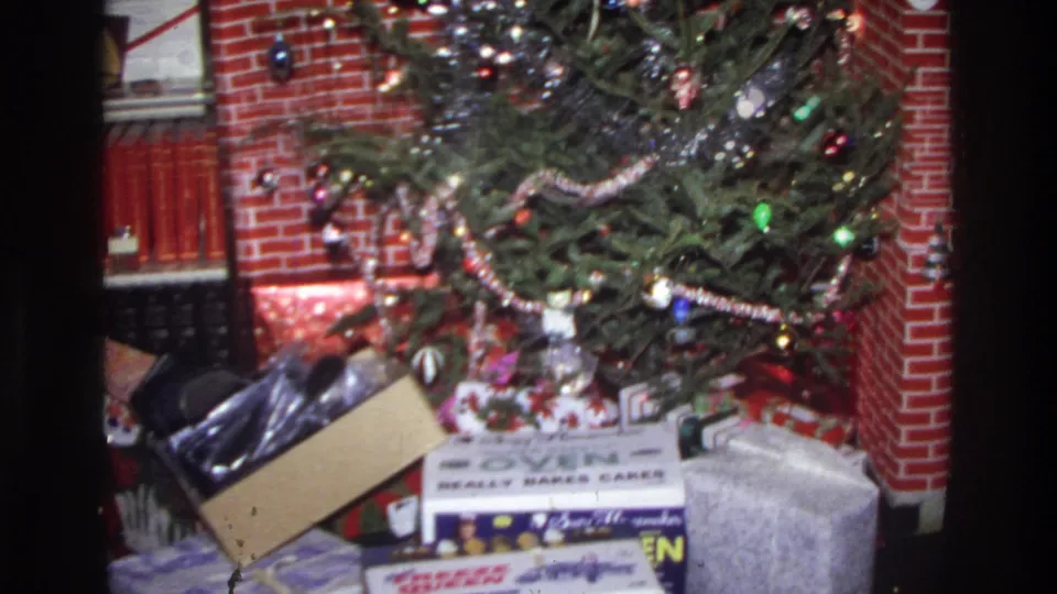 Closer view of a 1975 decorated Christmas tree with string lights and ornaments; wrapped presents at the base include a cardboard box with legible printed branding, set before a red brick fireplace.