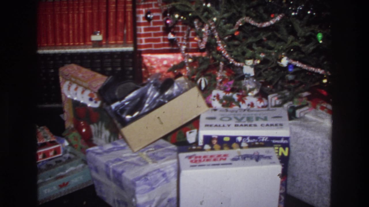 8mm home movie frame circa 1975 Washington DC: Christmas gifts under a tree, branded toy appliance boxes on a dark floor, brick fireplace surround behind.