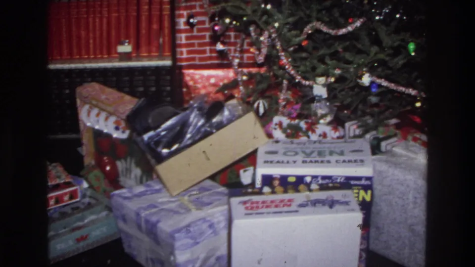 8mm home movie frame circa 1975 Washington DC: Christmas gifts under a tree, branded toy appliance boxes on a dark floor, brick fireplace surround behind.
