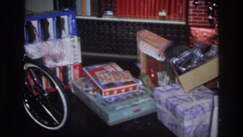 8mm home movie frame circa 1975 Washington DC: a purple bicycle wheel adjacent to wrapped gifts and board game boxes near a brick fireplace.