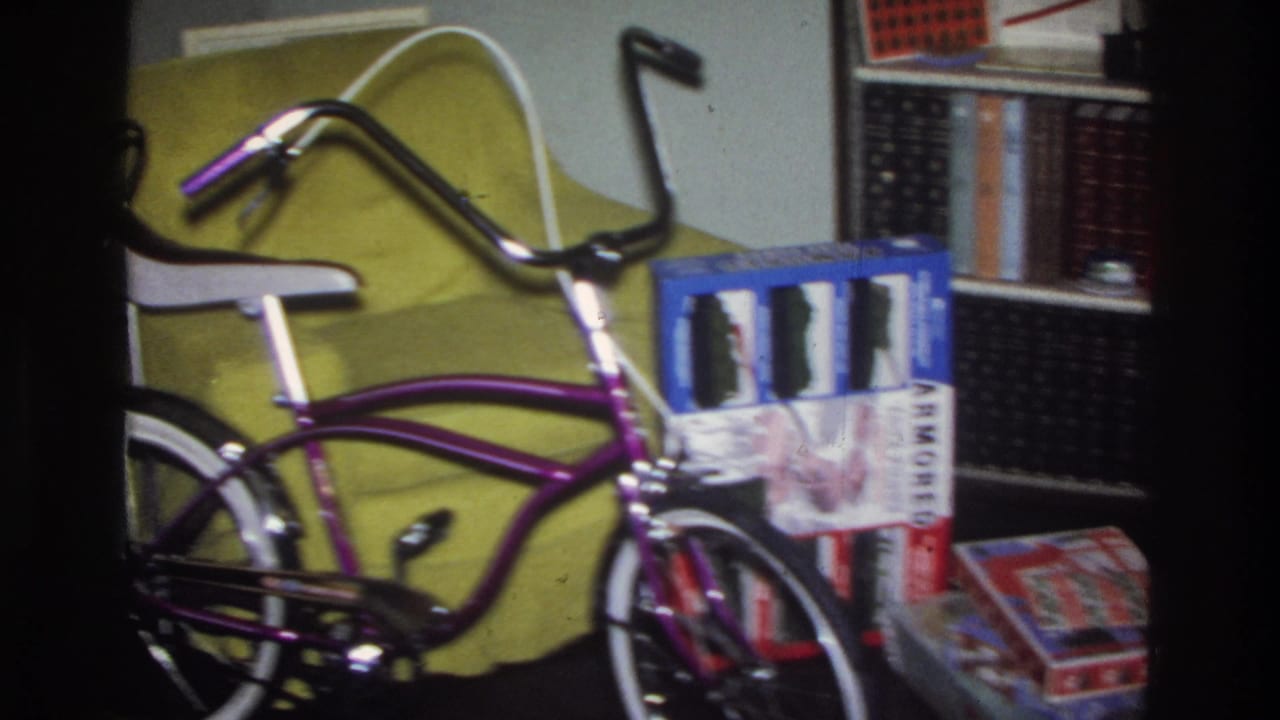 8mm home movie frame circa 1975 Washington DC: a purple banana-seat bicycle with chrome ape-hanger handlebars parked near a bookshelf and yellow upholstered furniture, gift boxes stacked nearby.