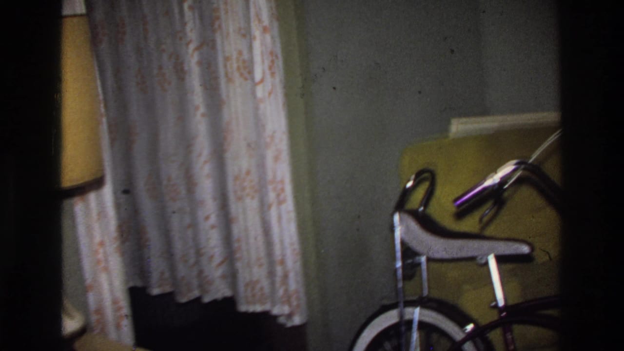 8mm home movie frame circa 1975 Washington DC: a domestic living room with patterned floral curtains, yellow upholstered furniture, and the purple bicycle partially in frame at right.
