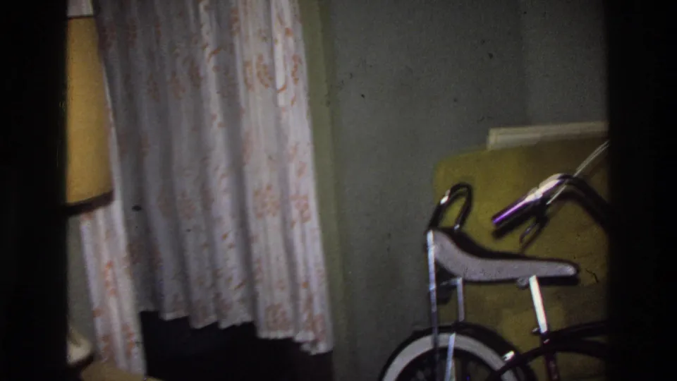 8mm home movie frame circa 1975 Washington DC: a domestic living room with patterned floral curtains, yellow upholstered furniture, and the purple bicycle partially in frame at right.
