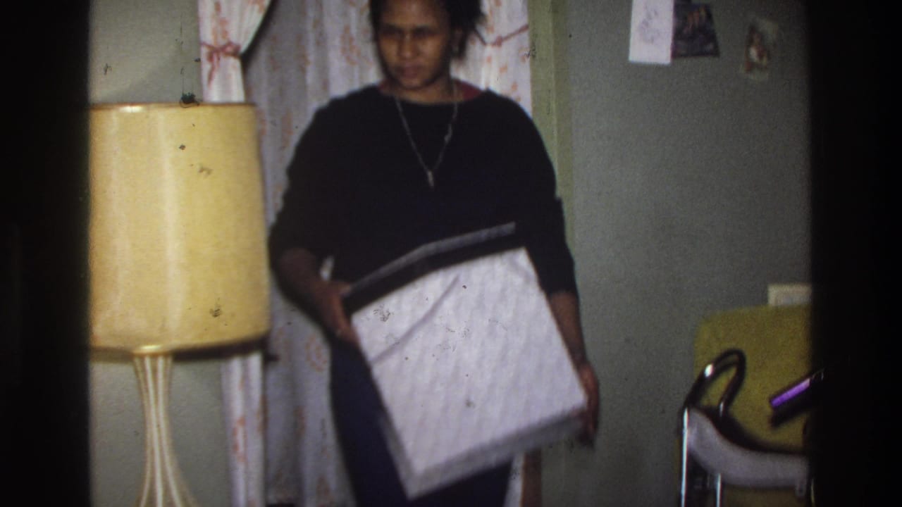 Black woman in black sweater holds a large patterned wig box in a home interior; yellow drum-shade lamp and patterned curtain visible; Washington DC, circa 1975.