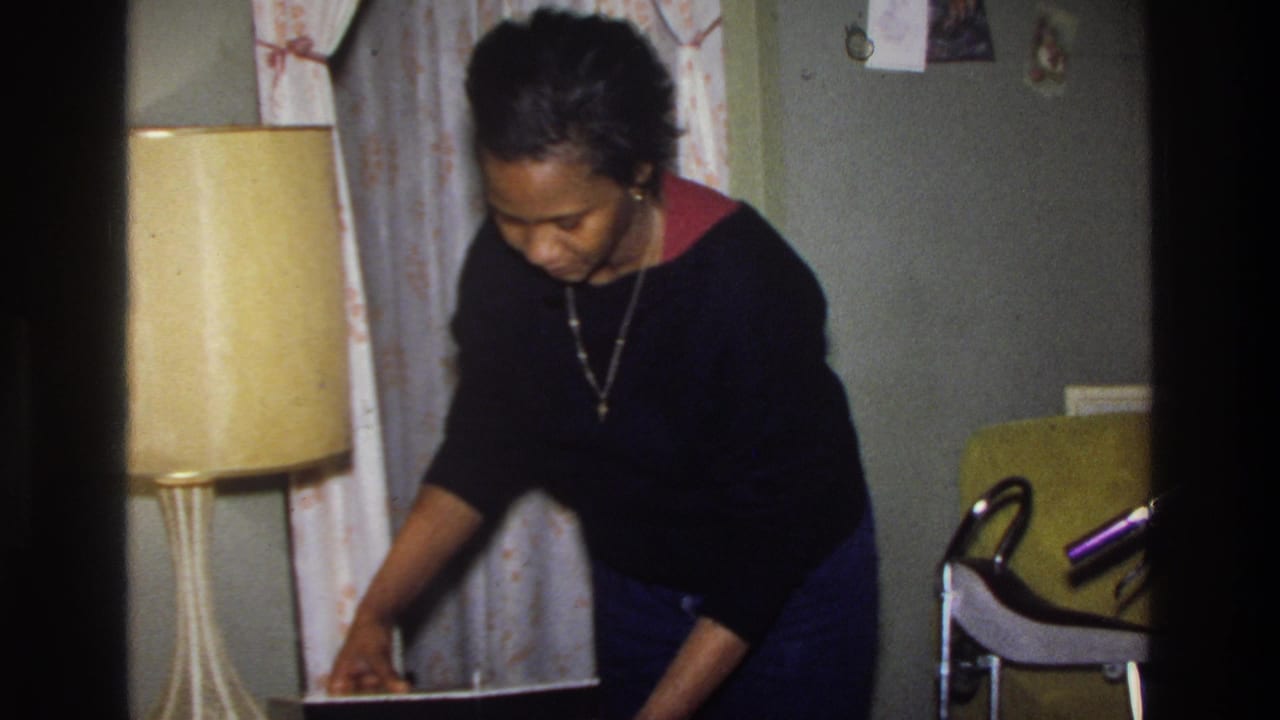 Woman leans over an open wig container on a surface, examining contents, with a yellow lamp and curtain visible behind her.