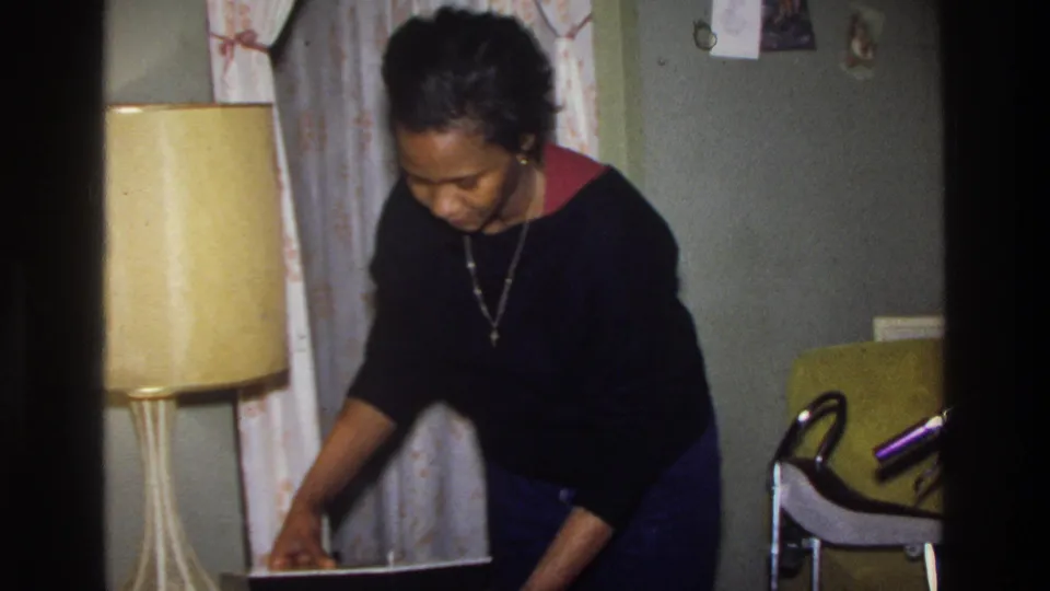 Black woman in black sweater bends over an open wig container in a home interior; yellow lamp and patterned curtain in background; Washington DC, circa 1975.