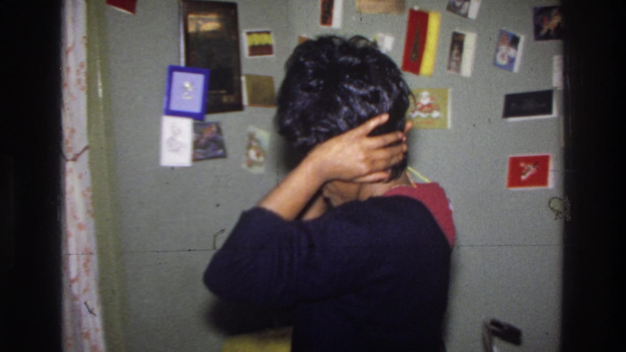 Black woman in black sweater adjusts a dark wig on her head, viewed from behind; wall densely pinned with greeting cards, postcards, and photographs; Washington DC, circa 1975.