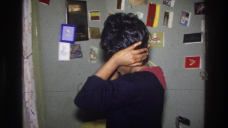 Black woman in black sweater adjusts a dark wig on her head, viewed from behind; wall densely pinned with greeting cards, postcards, and photographs; Washington DC, circa 1975.