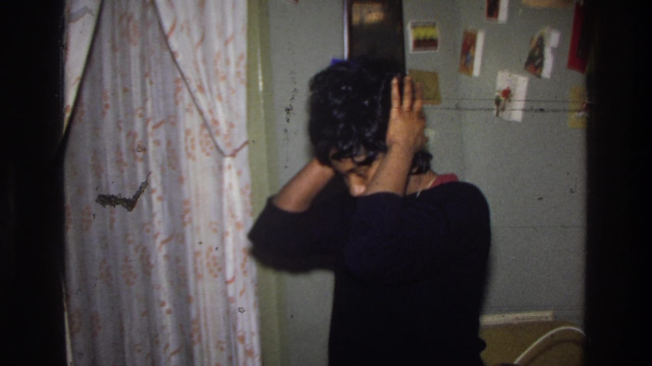 Black woman in black sweater smooths wig into place, viewed from side-rear; floral-patterned curtain and wall of pinned greeting cards visible; Washington DC, circa 1975.