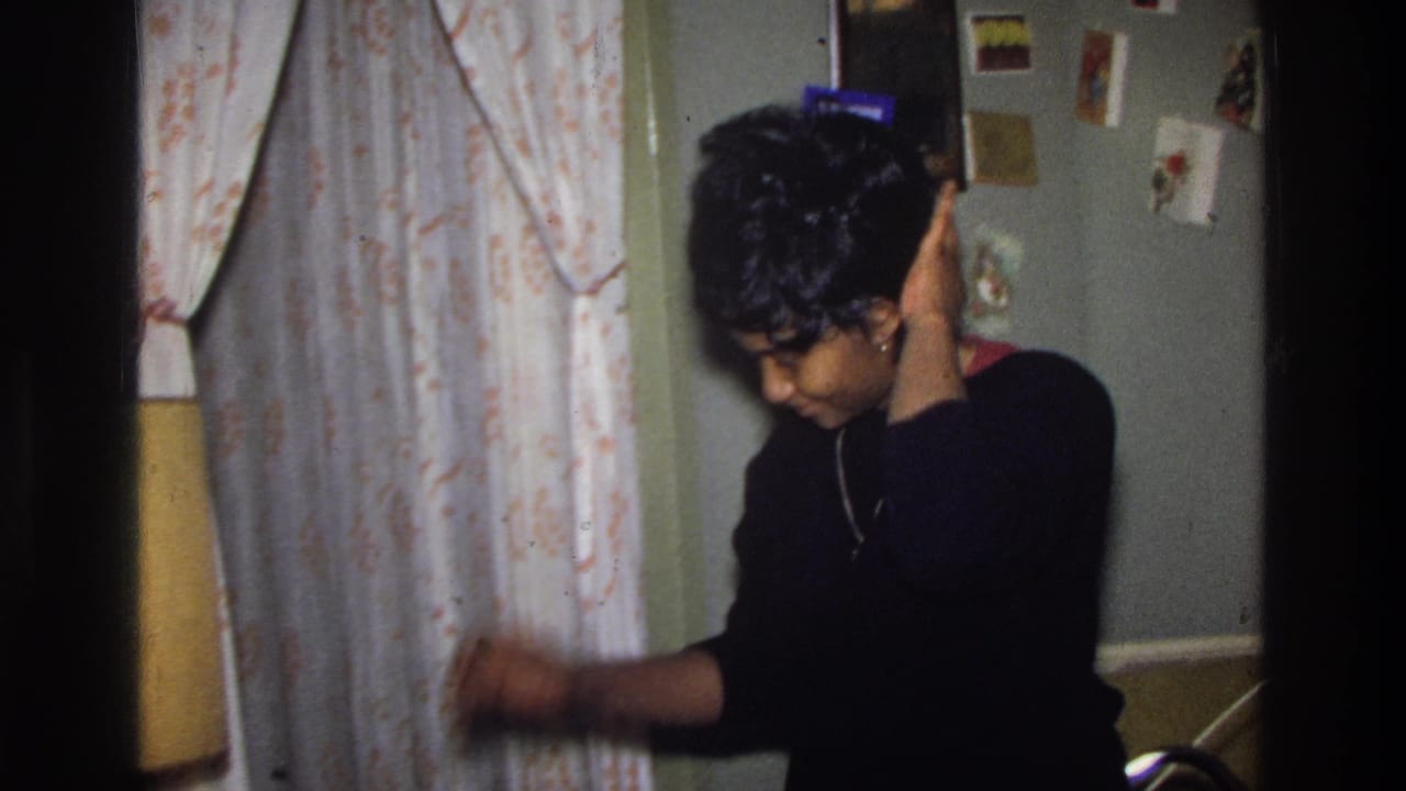 Woman in black sweater touches her hair in near-profile view, in a mid-1970s American living room with floral curtains and small cards pinned to a sage-green wall.
