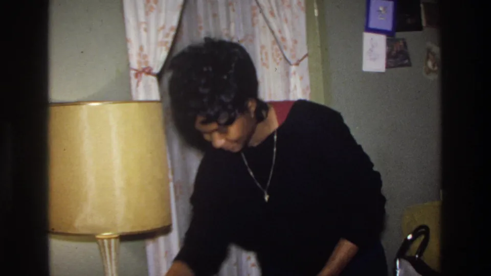 Woman in black sweater and cross pendant necklace stands beside a large amber drum lamp in a mid-1970s American living room; wall with pinned cards visible in background.