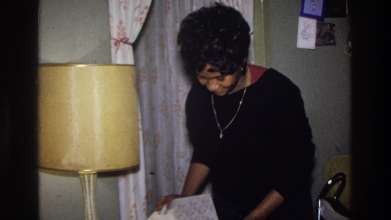 Woman in black sweater bends forward beside a large cylindrical amber lamp in a 1970s domestic living room, reaching or looking down toward a surface.