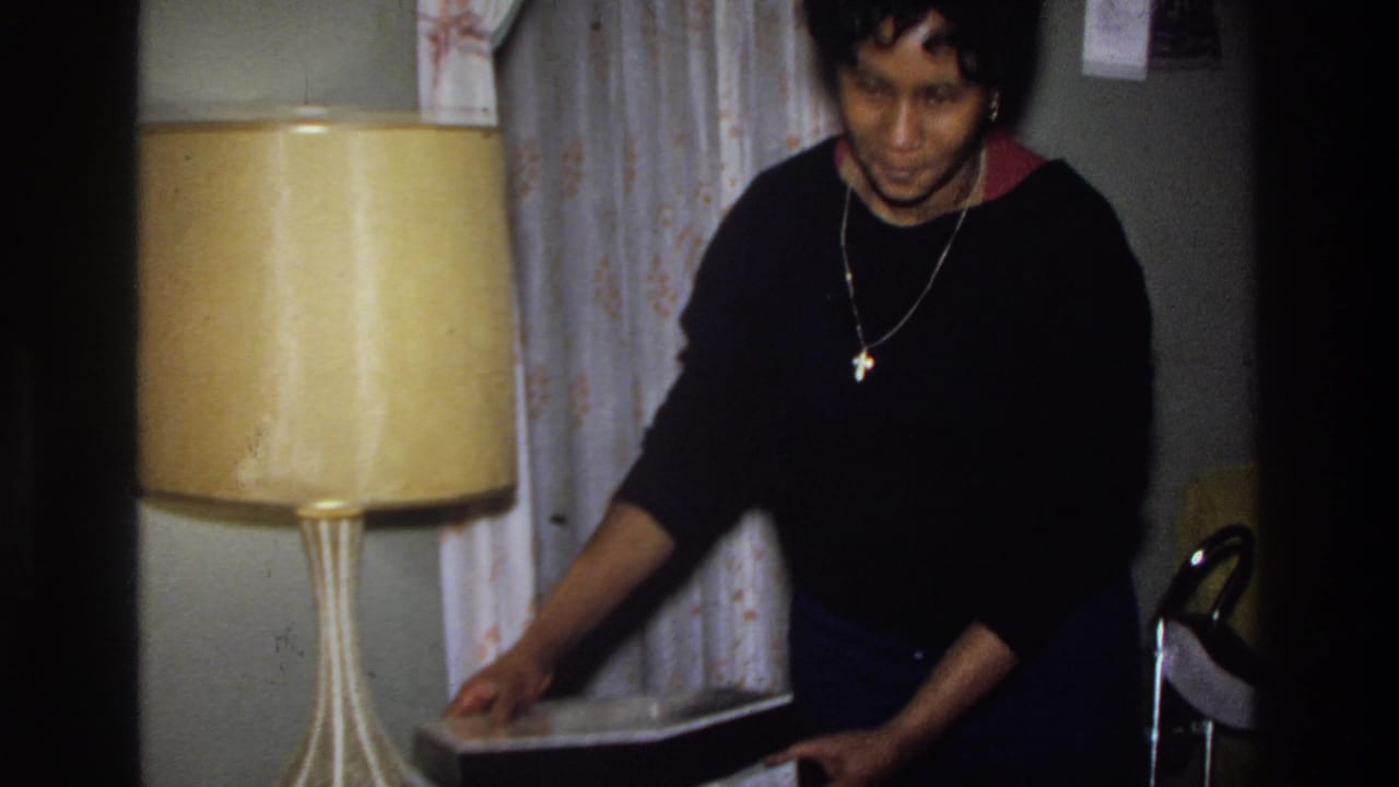 Woman in black sweater with visible cross pendant glances toward camera beside a large amber drum lamp in a mid-1970s American living room.