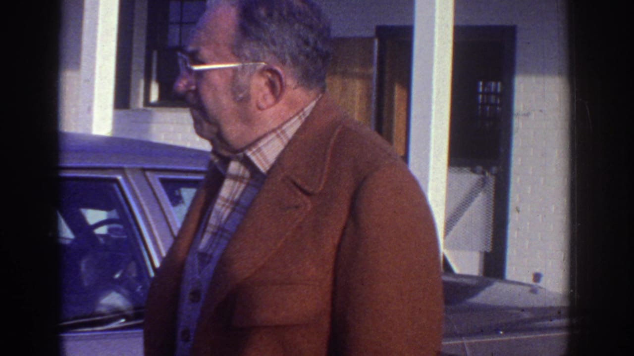 Man in rust-brown jacket and glasses walking past a dark parked car beside a white building.