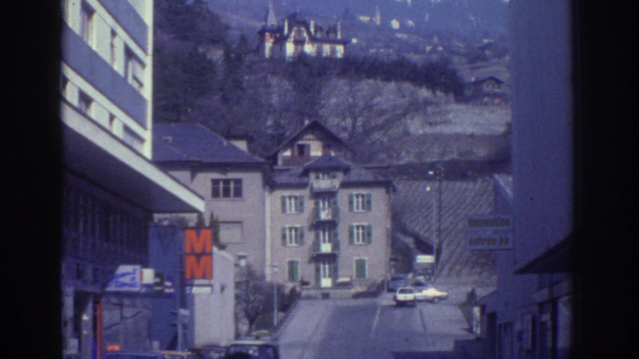 Street-level view in Bern, Switzerland, circa 1976, showing an orange M retail sign, period cars, mixed residential and commercial buildings, and a hillside backdrop with bare trees.