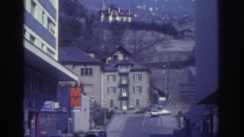 Street-level view in Bern, Switzerland, circa 1976, showing an orange M retail sign, period cars, mixed residential and commercial buildings, and a hillside backdrop with bare trees.