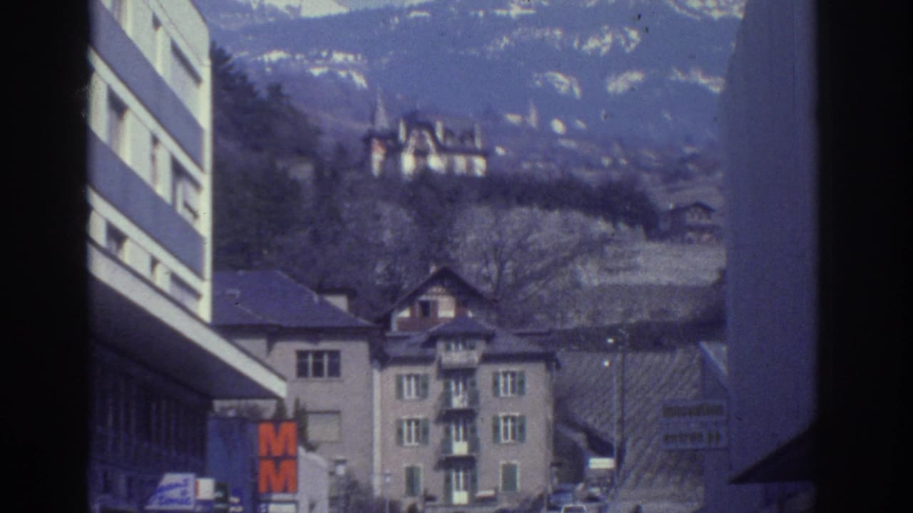 Continuing street pan in Bern, Switzerland, circa 1976, with the orange M retail sign, period automobiles, Swiss residential buildings, and wooded hillside visible.