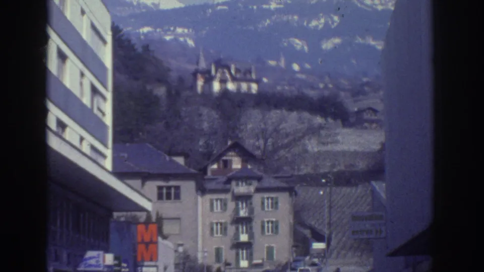Continuing street pan in Bern, Switzerland, circa 1976, with the orange M retail sign, period automobiles, Swiss residential buildings, and wooded hillside visible.