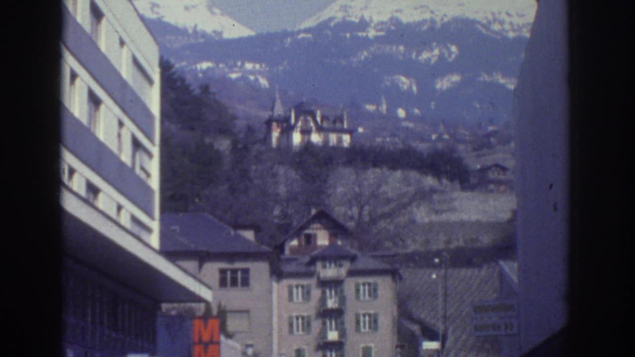 View past a modern concrete building edge toward snow-capped Alps, a turreted mansion on a wooded hillside, and mid-century residential blocks below.