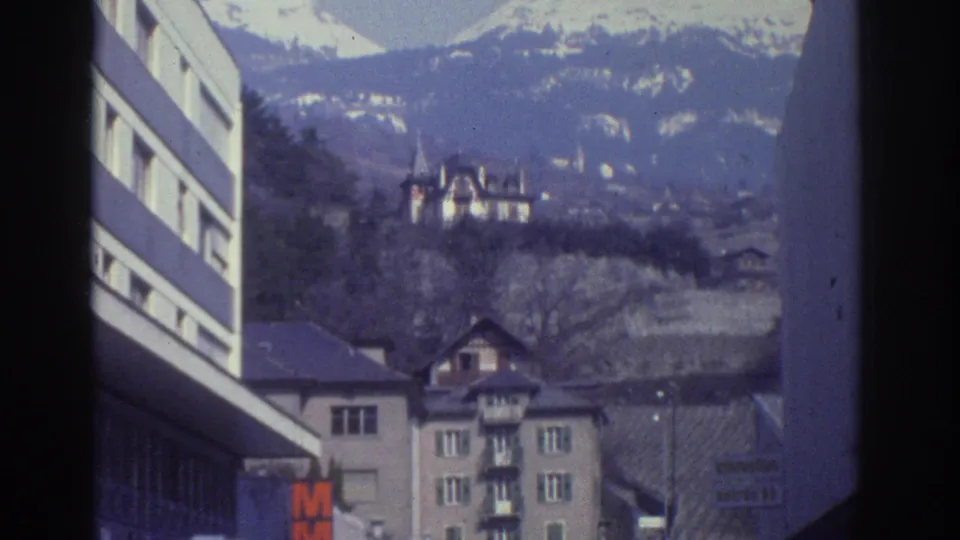 8mm frame from Bern, Switzerland, circa 1976: snow-capped mountains seen over a hillside with a turreted chateau and 1970s apartment buildings; partial orange commercial sign at left.