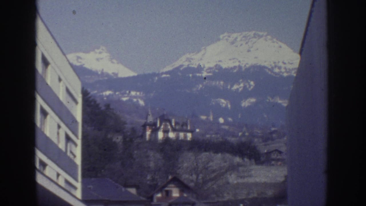 8mm frame from Bern, Switzerland, circa 1976: snow-capped Alps dominate the background as a turreted chateau sits among bare trees on the hillside; modern building at left edge.