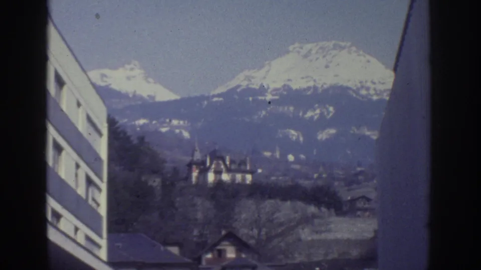 8mm frame from Bern, Switzerland, circa 1976: snow-capped Alps dominate the background as a turreted chateau sits among bare trees on the hillside; modern building at left edge.
