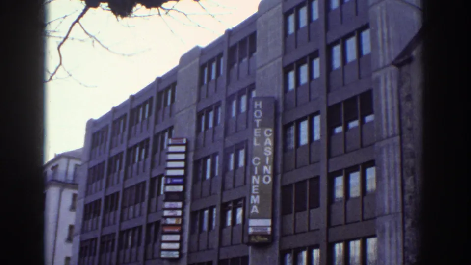 1970s Bern, Switzerland: brutalist multi-story building with vertical HOTEL CASINO CINEMA sign, repeating window pattern, bare winter tree branches, 8mm color cast.