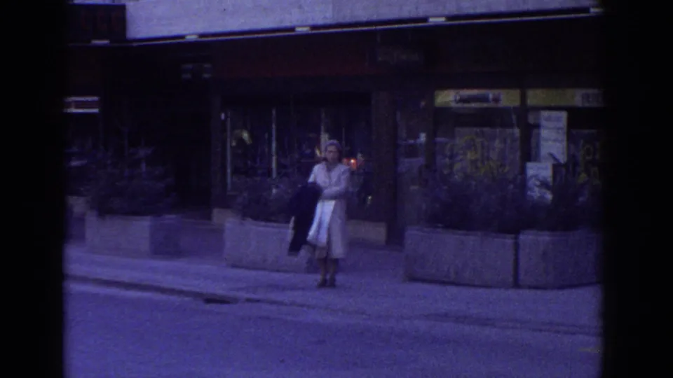 1976 Bern, Switzerland: woman in 1970s coat and skirt on a sidewalk beside raised concrete planters with greenery; commercial building facade with yellow signage at right.
