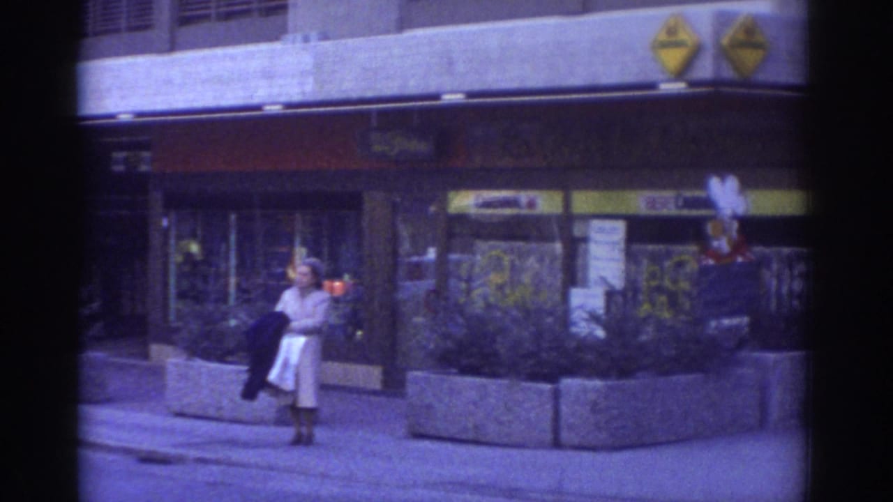 1976 Bern street scene: woman in period coat near concrete planter, yellow diamond commercial logos and red-yellow shop signage on building facade behind her, strong blue-purple 8mm color cast.