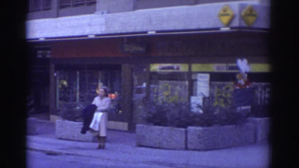 1976 Bern street scene: woman in period coat near concrete planter, yellow diamond commercial logos and red-yellow shop signage on building facade behind her, strong blue-purple 8mm color cast.