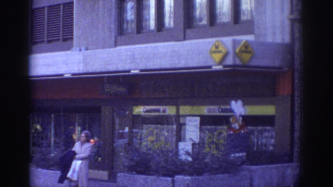 1976 Bern: woman in 1970s attire beside a sidewalk planter, yellow diamond logos and storefront signage on facade, strong 8mm blue-purple color cast throughout.