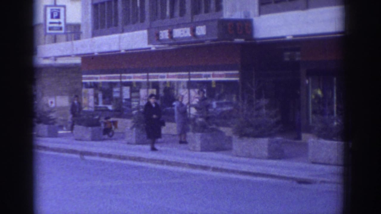 1976 Bern, Switzerland: pedestrians in dark coats pass storefronts and concrete planters; a moped or bicycle is visible in the mid-ground, captured on 8mm film with blue-cool tint.