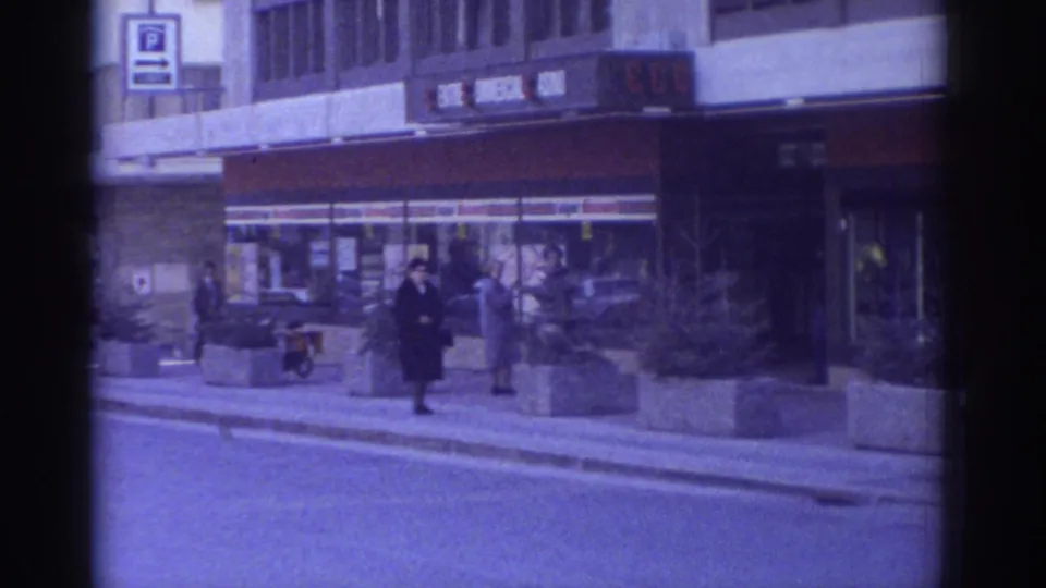 1976 Bern, Switzerland: pedestrians in dark coats pass storefronts and concrete planters; a moped or bicycle is visible in the mid-ground, captured on 8mm film with blue-cool tint.