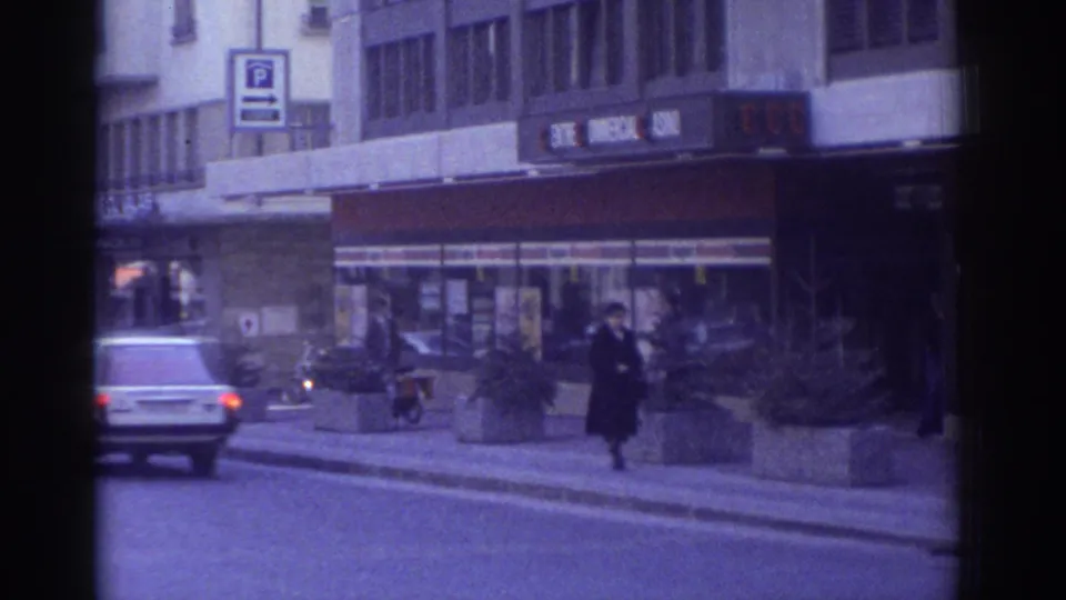 1976 Bern, Switzerland: a light-colored automobile moves along the street as a pedestrian crosses the pavement; a blue 'P' parking sign is prominent, shot on 8mm film.