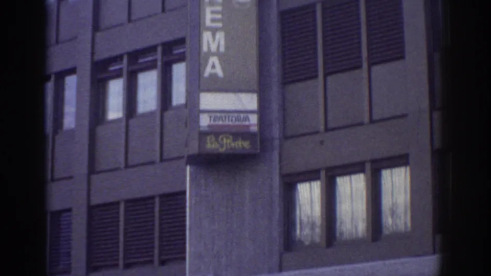 CINEMA sign above Trattoria restaurant signage on a modernist building facade, Bern, Switzerland, circa 1976, aged 8mm film with purple-blue color shift.