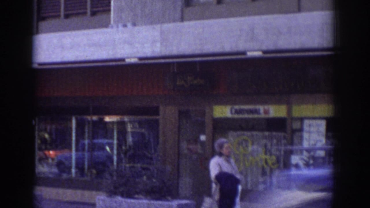 Woman walking past a storefront with a Cardinal sign; blue color cast from aged 8mm film.