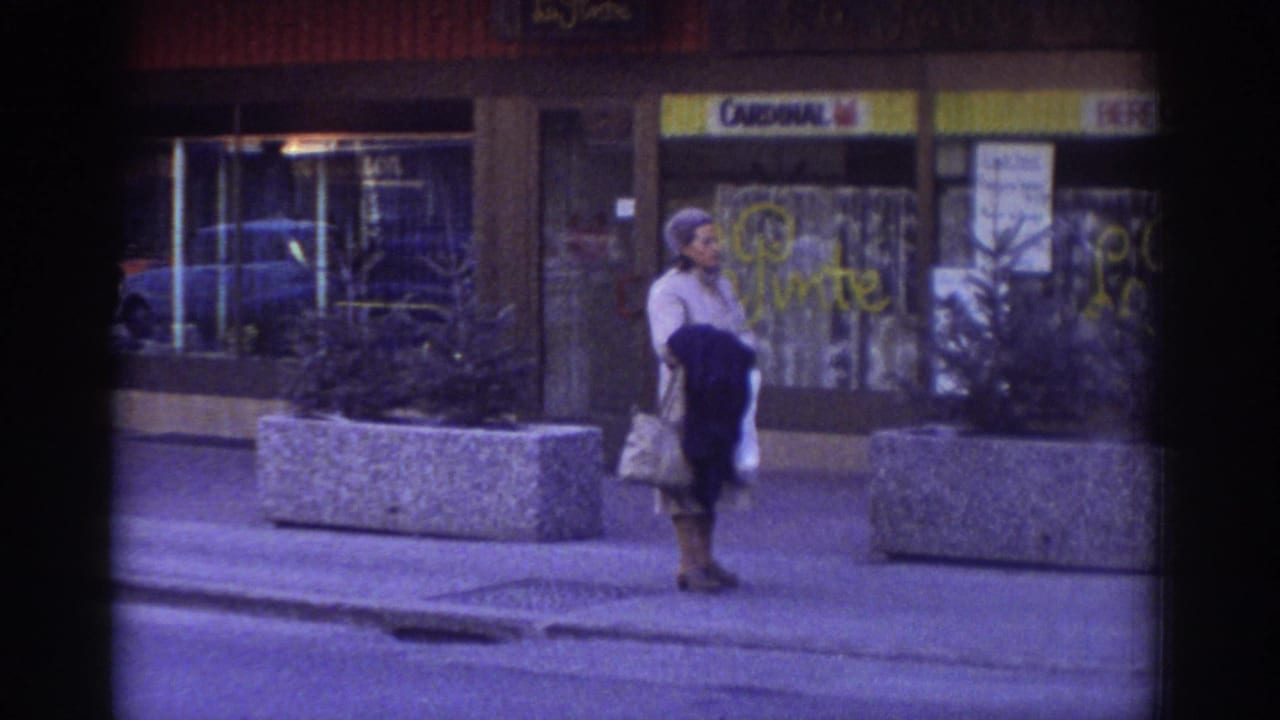 Woman carrying bags pauses near a Cardinal-signed shopfront in what appears to be Bern, Switzerland, c.1976; concrete planter and graffiti on glass visible.