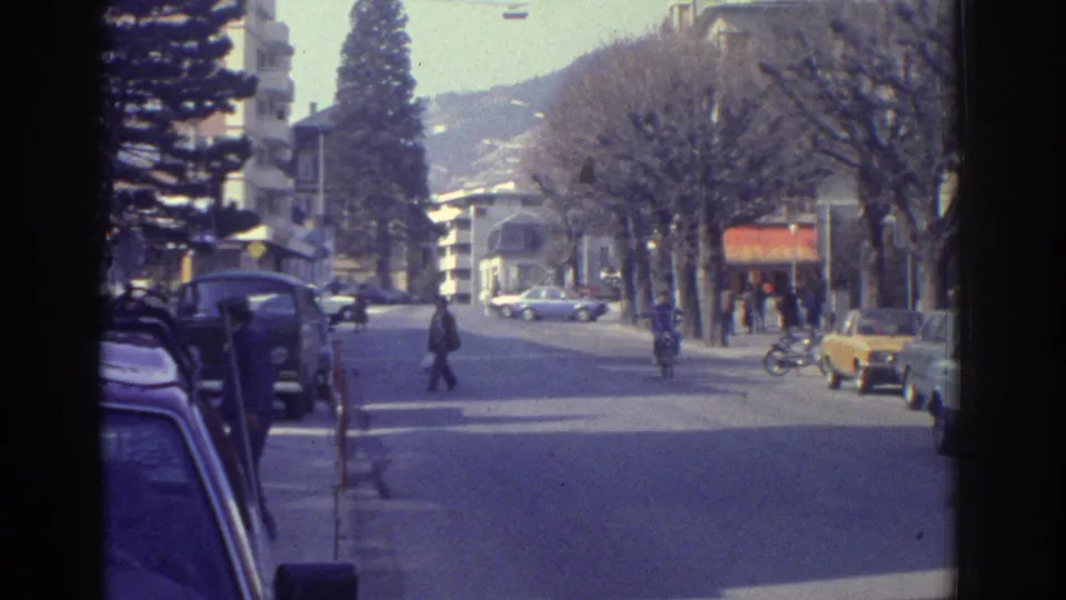 Tree-lined boulevard in what appears to be Bern, Switzerland, c.1976; 1970s-era sedans at curb, cyclist crossing, bare deciduous trees, hills in distance.