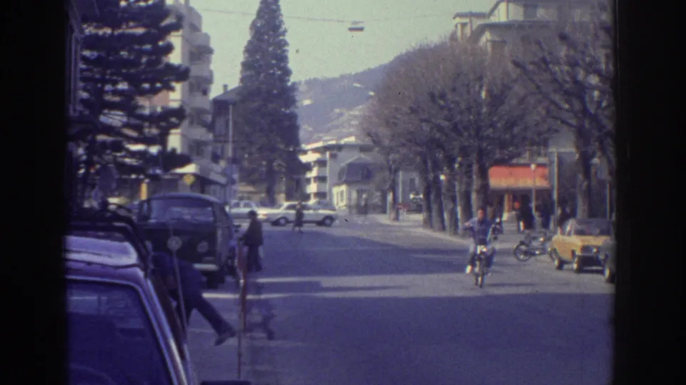 Street-level view of a tree-lined avenue in what appears to be Bern, Switzerland, c.1976; yellow car in foreground, motorcyclist, parked sedans, and low hills in background.