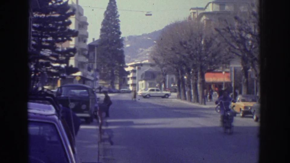 1976 8mm home movie of a Bern, Switzerland boulevard in winter with a tall evergreen conifer on the left, 1970s cars, a motorcyclist, and pedestrians.