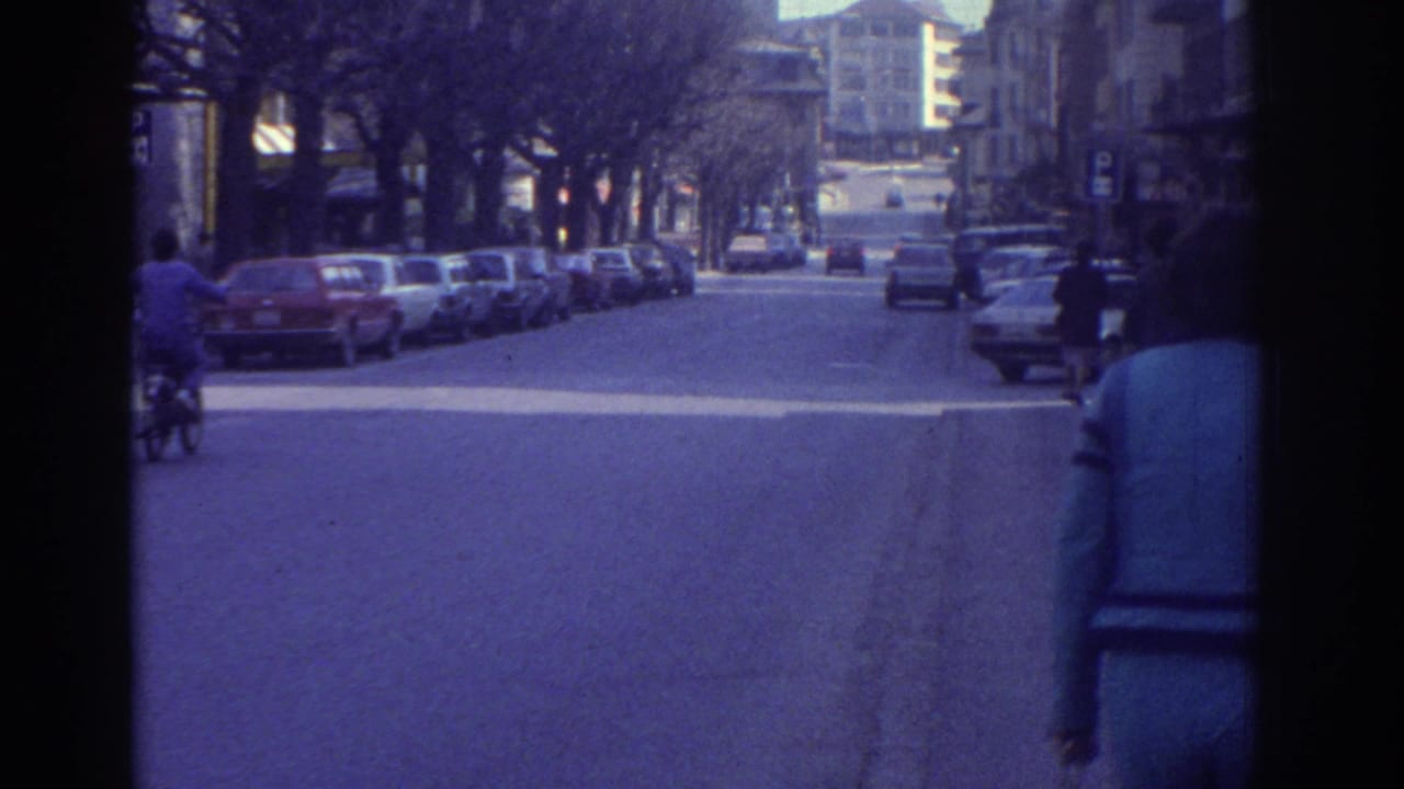 1976 8mm footage of a Bern, Switzerland boulevard showing a cyclist in the road, parked 1970s saloon cars, bare winter trees, and multi-story buildings.