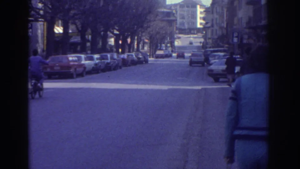 1976 8mm footage of a Bern, Switzerland boulevard showing a cyclist in the road, parked 1970s saloon cars, bare winter trees, and multi-story buildings.