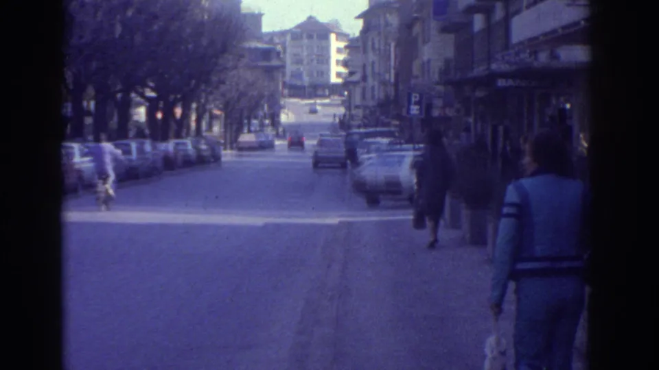 1976 8mm home movie showing a pedestrian in blue clothing walking along a Bern city street past shop awnings with partial signage, parked 1970s cars, and a traffic light.