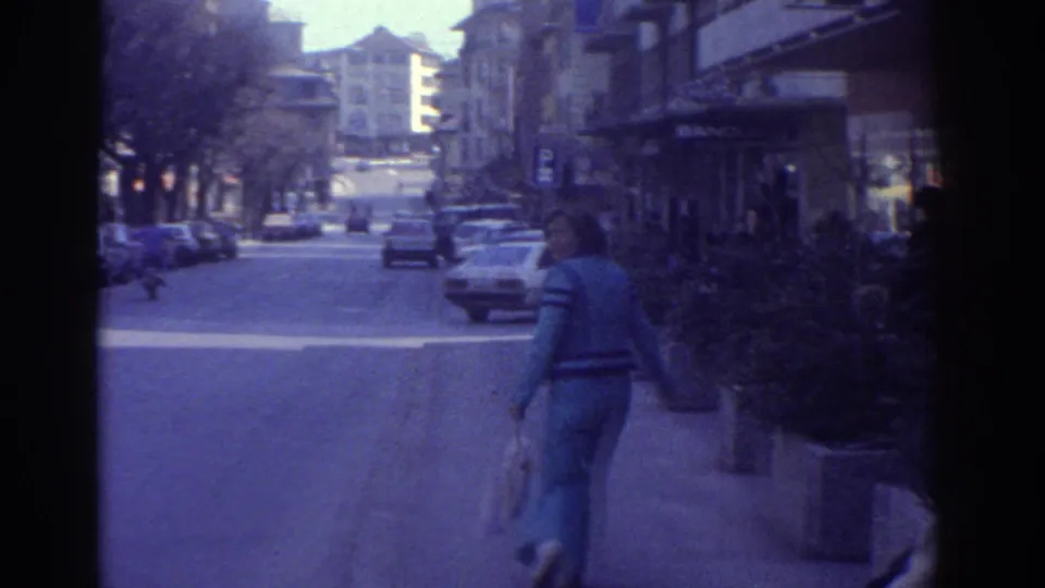1976 8mm home movie of a Bern city street showing a pedestrian in blue clothing, shop awnings, parked 1970s cars, a traffic light, and a small potted conifer in the foreground.