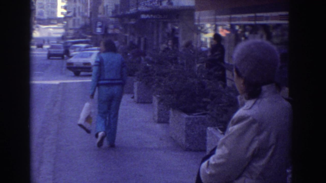 Person in blue jacket and trousers walking away on a city sidewalk; a second figure in a knit hat stands in the foreground; parked cars and concrete planters visible.