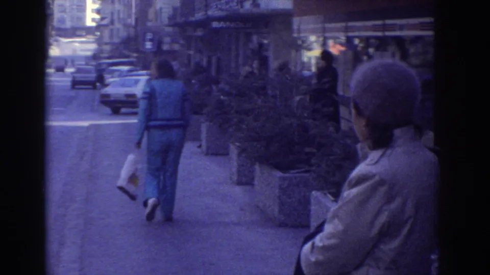 1976 Bern, Switzerland on 8mm film: blue-clad pedestrian walking away from camera, bystander in beanie hat in foreground, 1970s European cars parked along a commercial sidewalk.