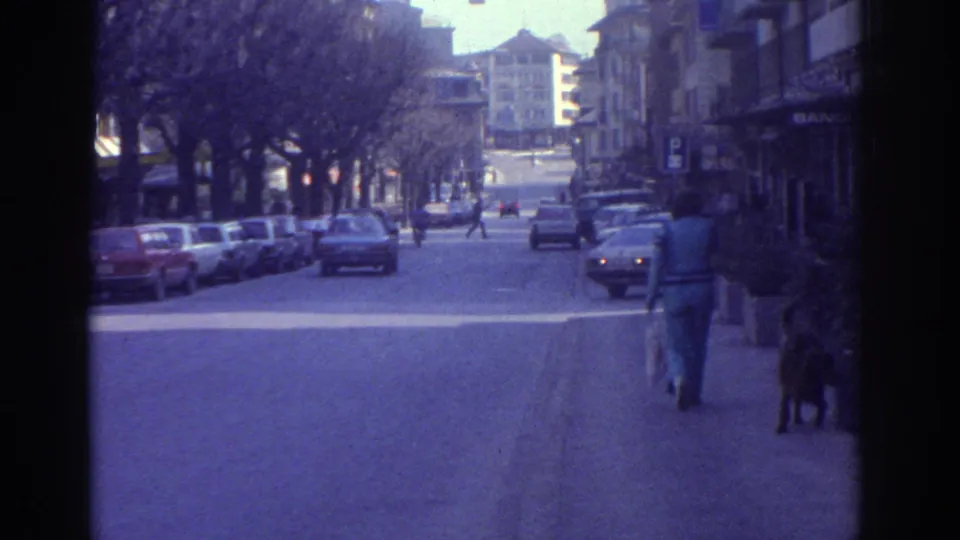 1976 Bern, Switzerland on 8mm: wide commercial street with period European cars, bare winter trees, multi-story buildings, pedestrian with a dog, and partial sign reading 'BAN-'.